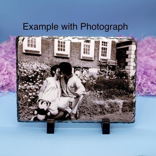 A rectangular slate photo plaque on a black display stand, showing a black-and-white photograph of a couple in front of a brick house. The image has the text 'Example with Photograph' at the top and is set against a blue background with pink decorative elements.