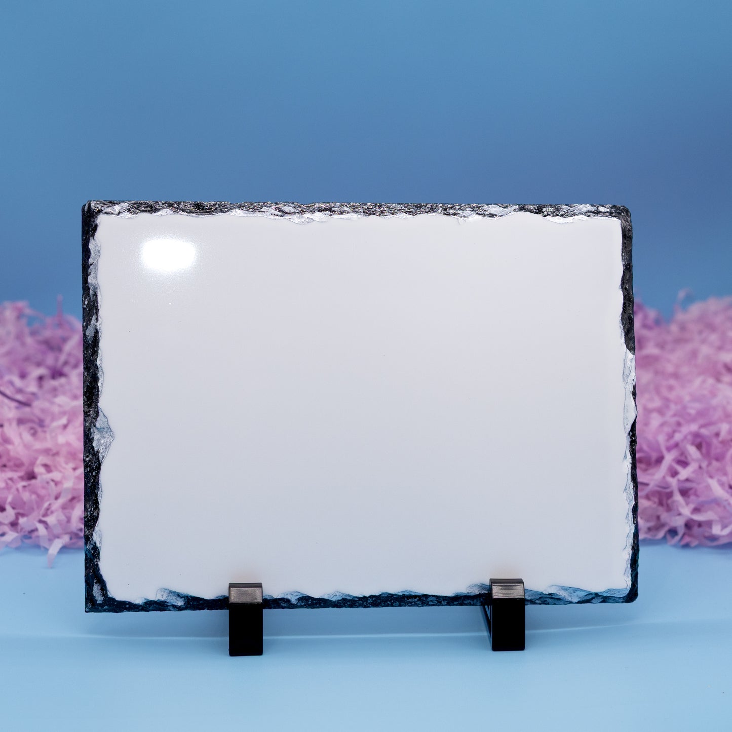 A rectangular slate with rough dark natural edges, displayed on a small black stand against a blue background with pink shredded paper accents.