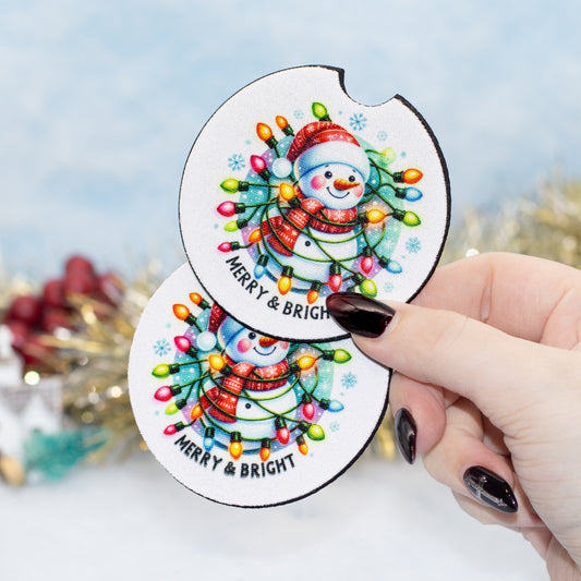 A hand holds two round white coasters featuring a colourful Christmas snowman design with glowing holiday lights and the text 'MERRY & BRIGHT'. The background is softly blurred with festive decorations.