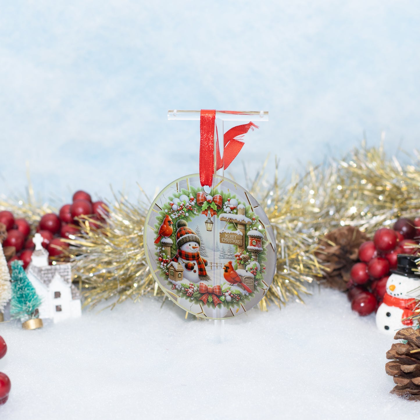 A round Christmas ornament displayed on a clear stand, featuring a festive winter scene with snowman, snowman figurines, red birds, and a wreath, hanging from a red ribbon. The ornament is styled on a white surface with gold tinsel, red berries, pinecones, and small decorative houses in the background.