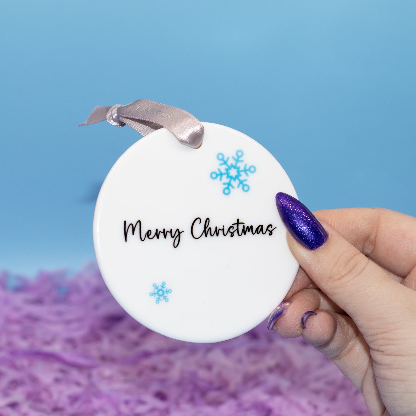 A hand holds a round white Christmas ornament that reads 'Merry Christmas' in black script, decorated with two light blue snowflakes, against a blue and purple background.