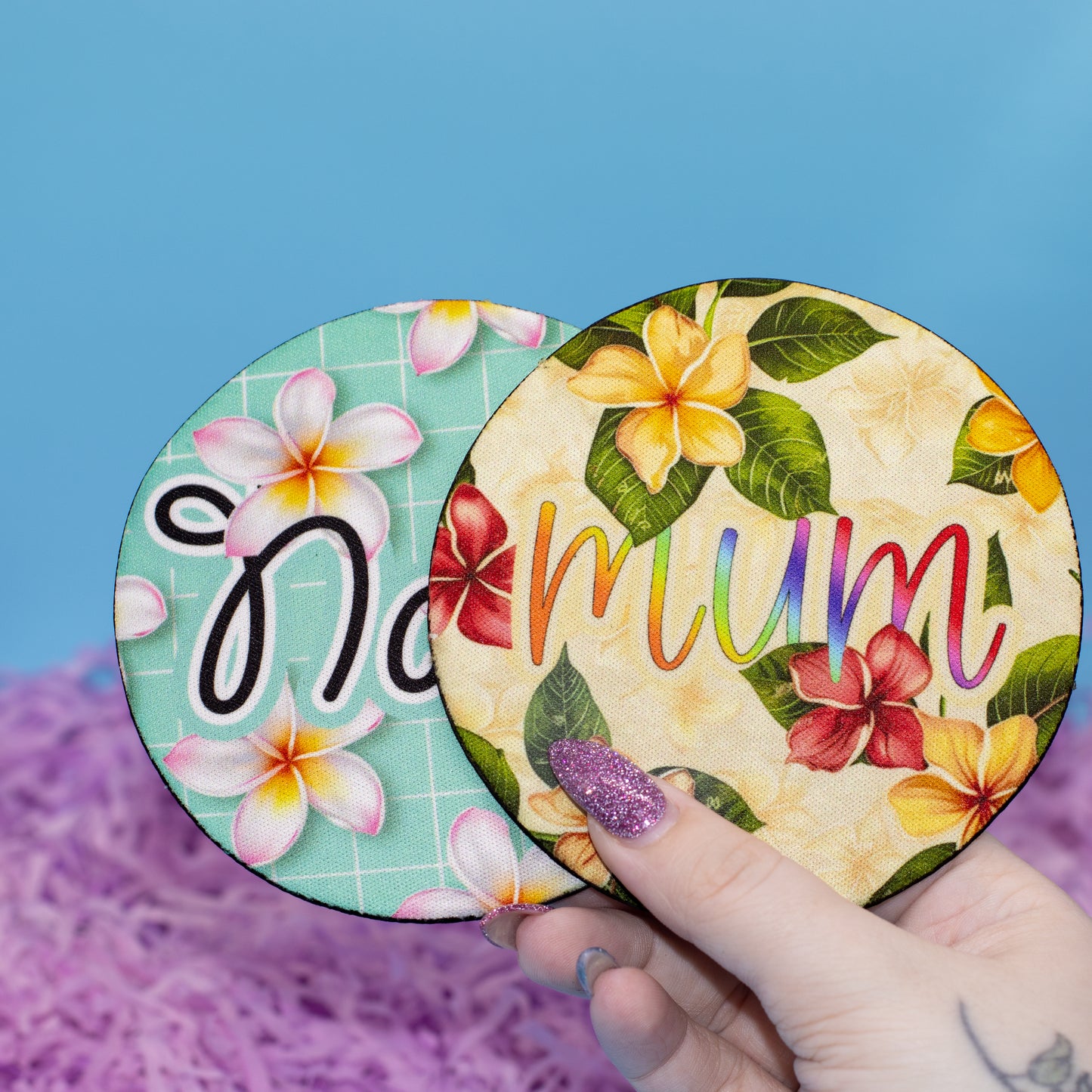 Two round coasters with tropical floral designs are shown, one partially covered by the other. A hand with glittery purple nail polish holds them against a blue background with purple shredded paper at the bottom.