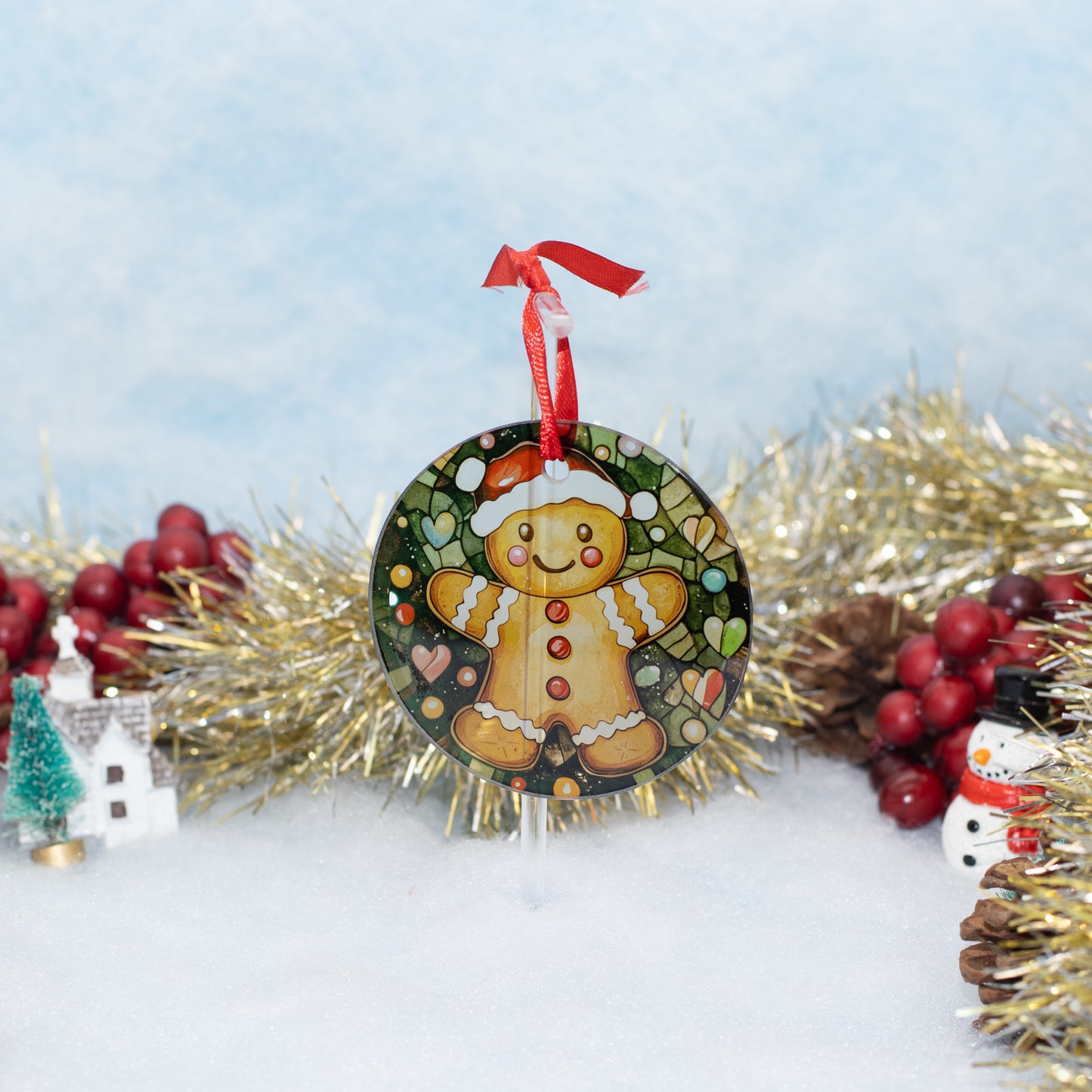 A round Christmas ornament featuring a gingerbread man on a dark green background, hanging from a red ribbon. The ornament is styled on white faux snow with gold tinsel, red berries, pinecones, and a small snowman figurine against a soft light blue backdrop.