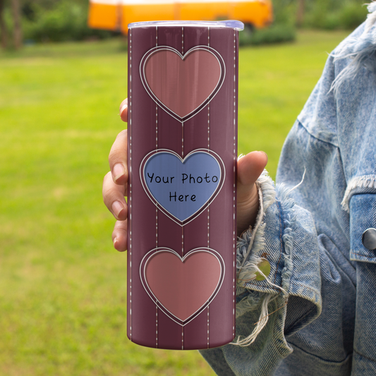 A person holding a personalized stainless steel tumbler outdoors. The tumbler has a dark red-burgundy background with three heart-shaped photo frames outlined in white stitching, including a central blue heart with the text 'Your Photo Here'.