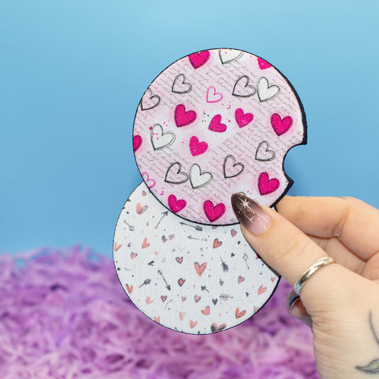A hand holds two round car coasters with heart prints against a blue background with purple shredded paper at the bottom.