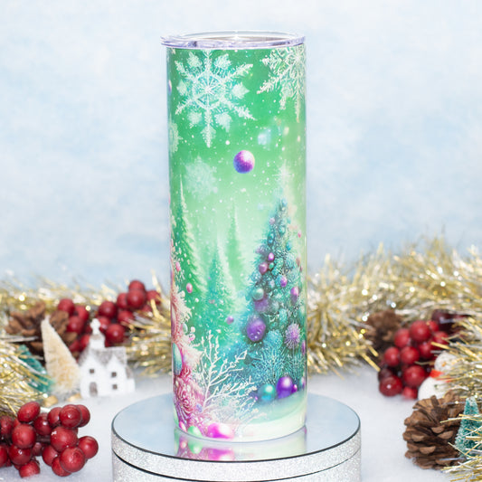 A tall, slim tumbler with a clear lid, decorated with a festive Christmas print featuring a green gradient background, snowflakes, evergreen trees, and purple ornaments. The tumbler is displayed on a round silver mirror stand, surrounded by holiday decorations like red berries, pinecones, and gold tinsel against a soft, light blue background.