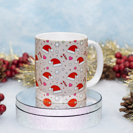 A white ceramic mug with a festive Christmas pattern of Santa hats, candy canes, hearts, and stars on a light beige background, displayed on a round silver glitter tray.