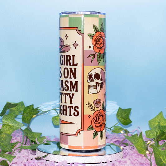 A tall insulated tumbler with a retro checkerboard design in pastel pink, green, and cream, featuring illustrations of a skull, roses, and a cowboy hat. The tumbler is displayed on a mirrored pedestal surrounded by purple flower petals and green ivy against a light blue background.