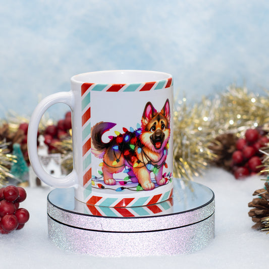 A white ceramic mug featuring a colorful illustration of a German Shepherd dog wrapped in festive Christmas lights, displayed on a round silver glitter pedestal with holiday decorations in the background.