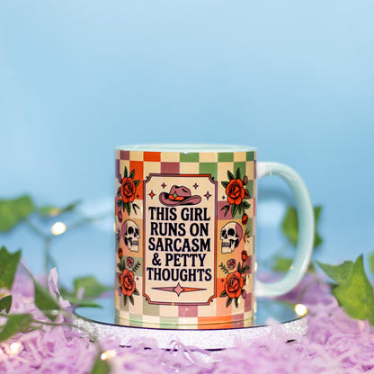 A ceramic mug with a colorful retro checkered border and a central graphic featuring a cowboy hat, skulls, roses, and the text 'THIS GIRL RUNS ON SARCASM & PETTY THOUGHTS'. The mug is displayed on a silver glittery stand surrounded by purple shredded paper, green leaves, and fairy lights against a soft blue background.