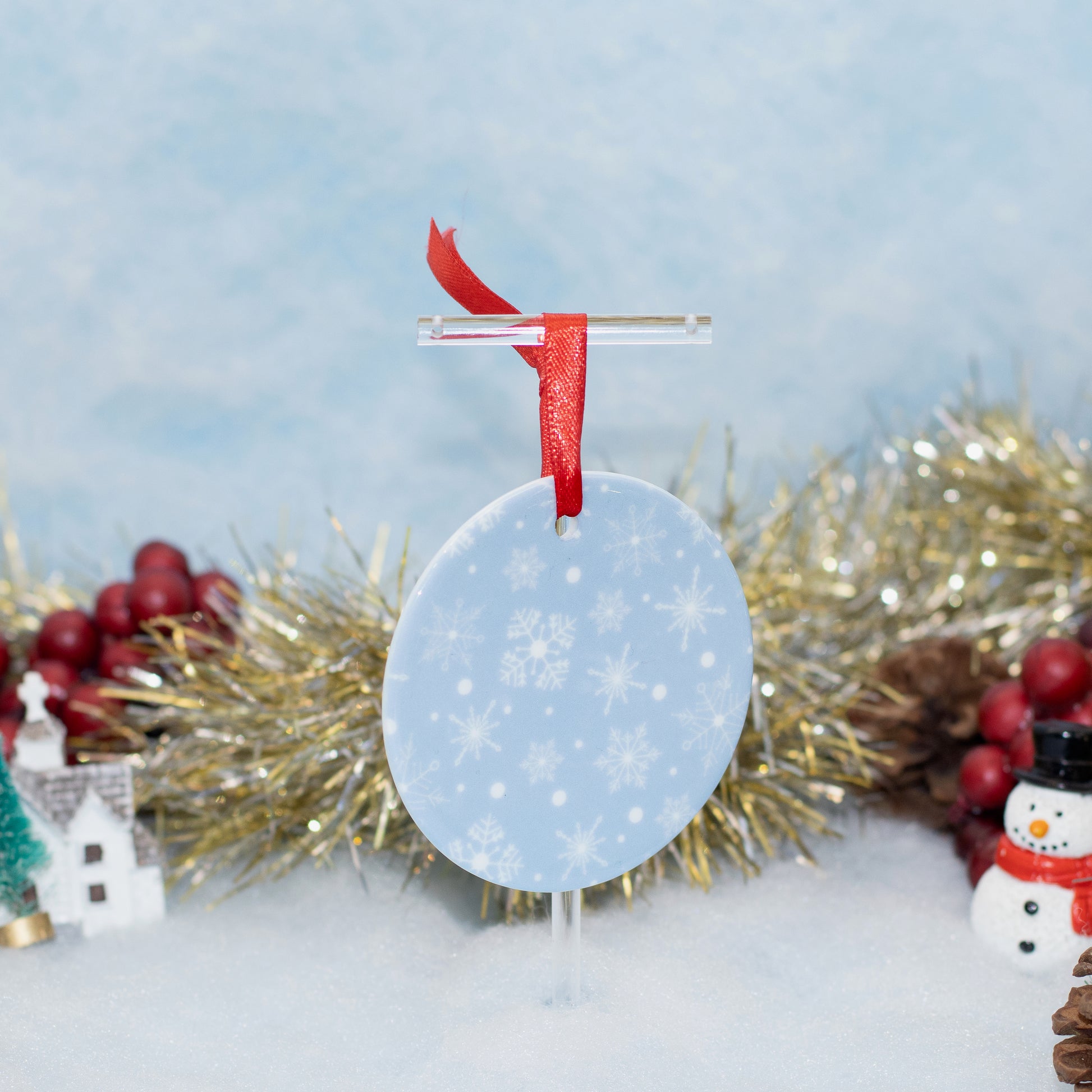 A round light blue Christmas ornament with white snowflakes, hanging from a clear stand with a red ribbon, set against a snowy winter scene with gold tinsel, red berries, and a small snowman decoration.