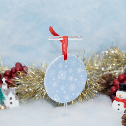 A round light blue Christmas ornament with white snowflakes, hanging from a clear stand with a red ribbon, set against a snowy winter scene with gold tinsel, red berries, and a small snowman decoration.