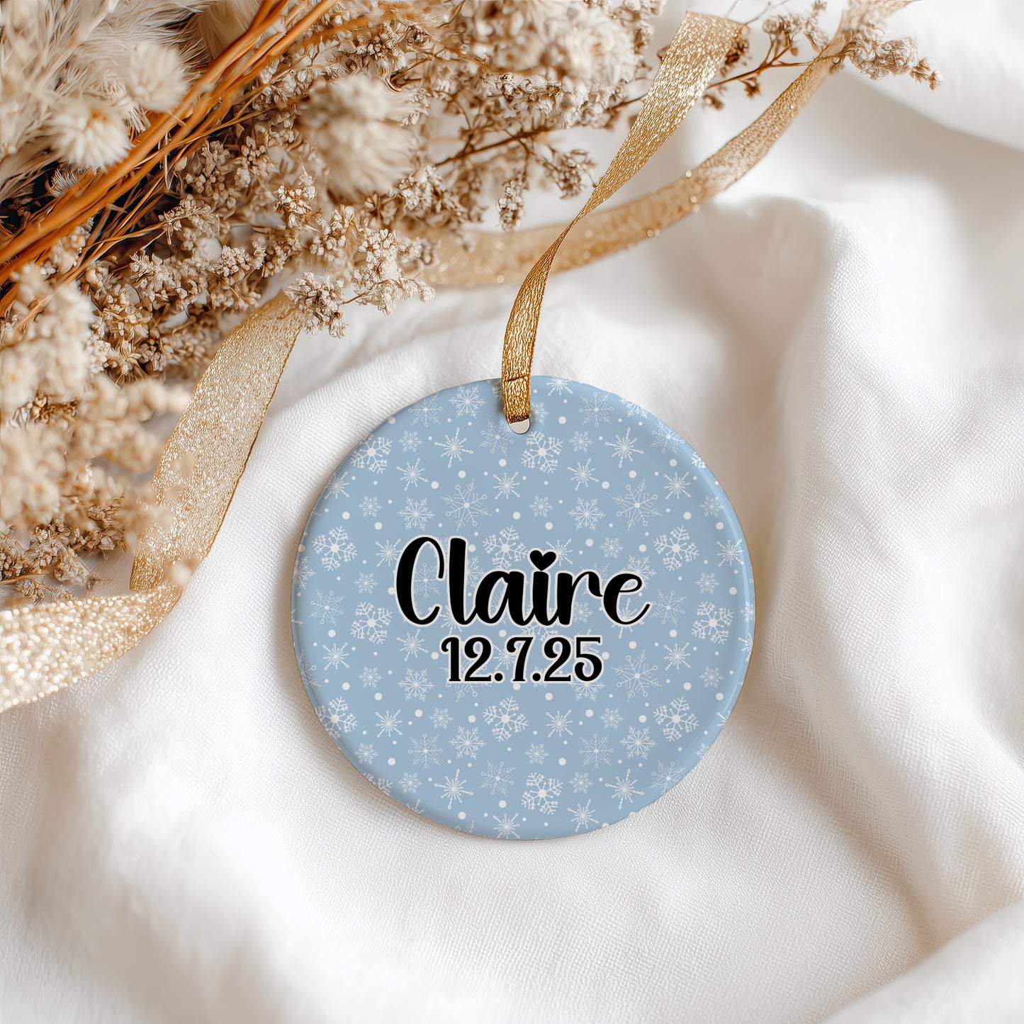 A round personalized Christmas ornament with a light blue snowflake pattern, hanging from a gold ribbon. The ornament is printed with the name 'Claire' and the date '12.