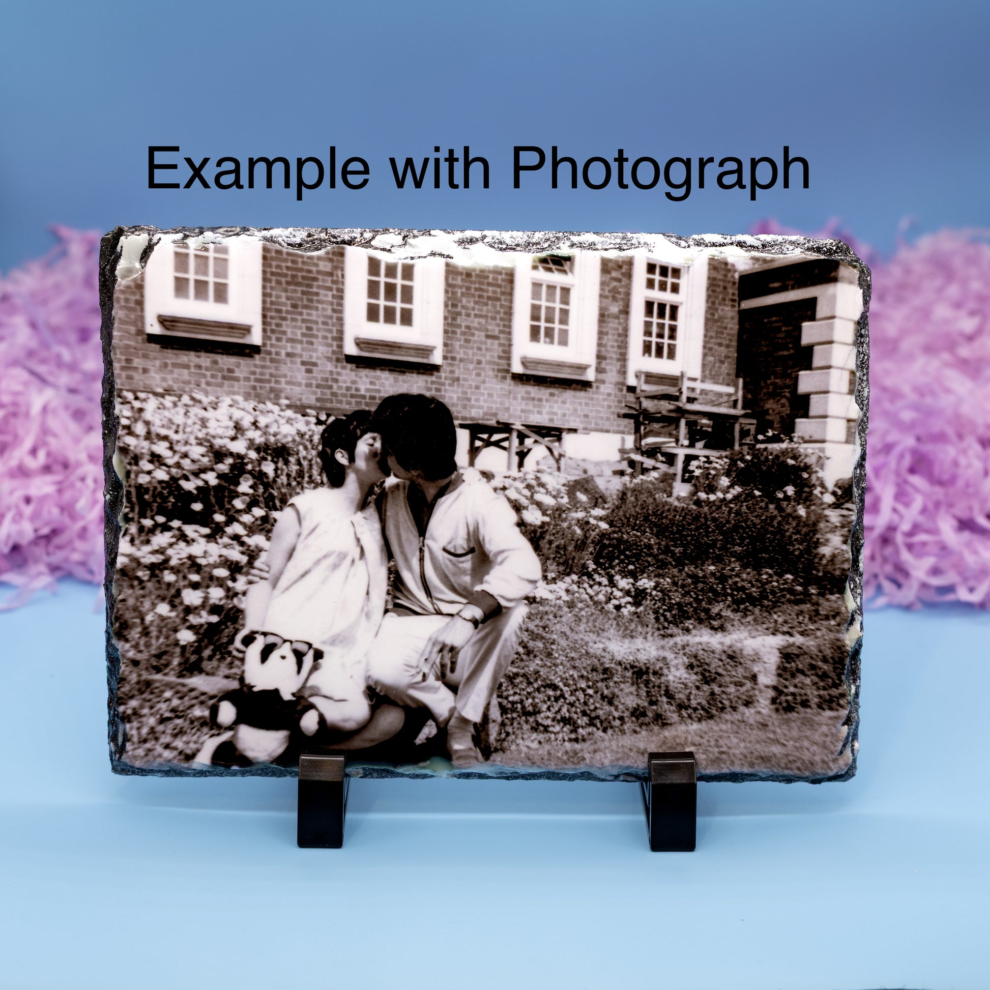 A rectangular slate photo plaque on a black display stand, showing a black-and-white photograph of a couple in front of a brick house. The image has the text 'Example with Photograph' at the top and is set against a blue background with pink decorative elements.