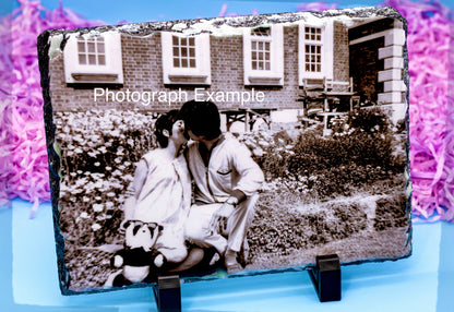 A rectangular slate photo plaque displaying a black-and-white photograph of a couple in front of a brick house, shown on a light blue surface with purple shredded paper in the background. A white text watermark reading 'Photograph Example' is overlaid across the center.