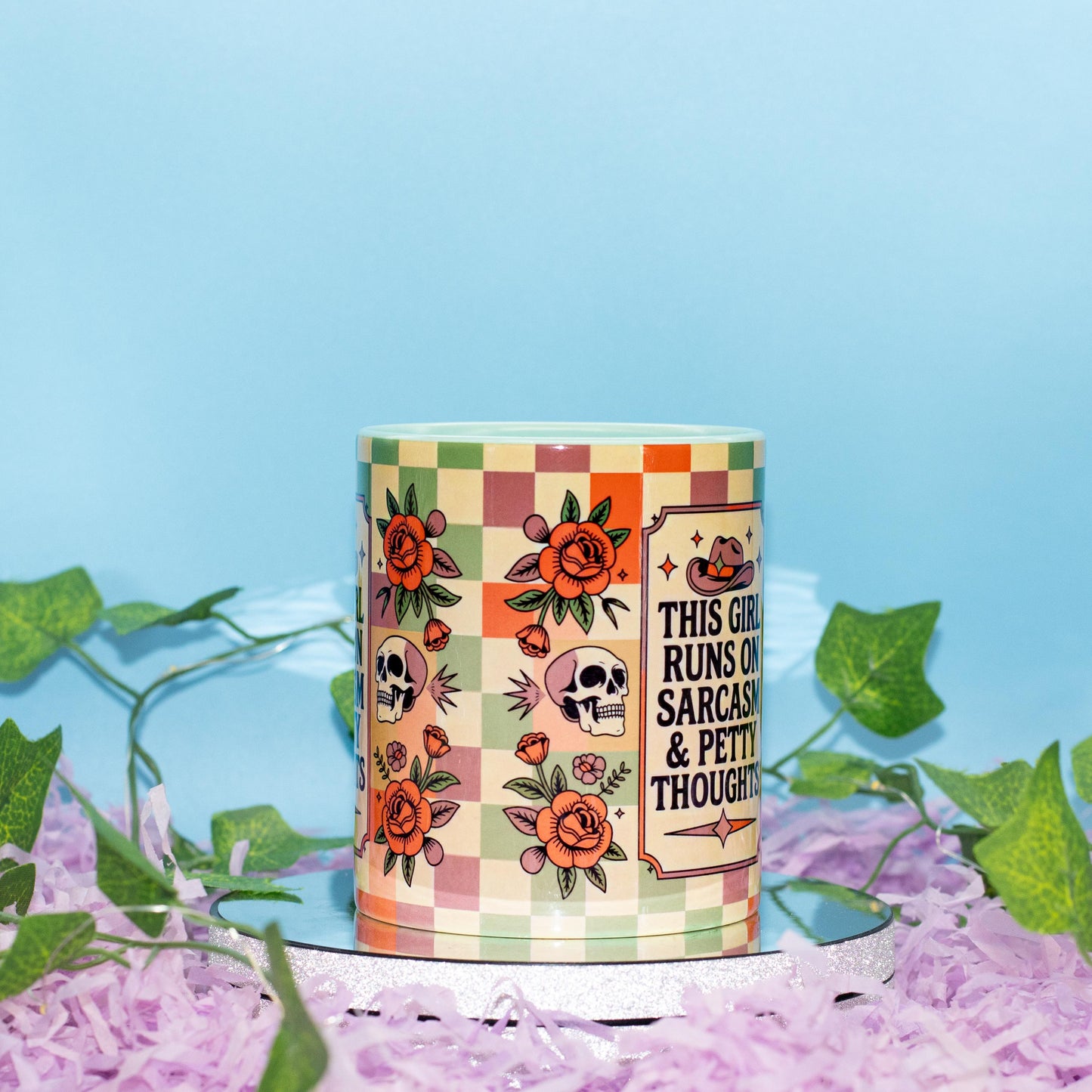 A cylindrical mug with a colorful retro checkerboard pattern and illustrations of skulls, roses, and pumpkins, featuring the text 'THIS GIRL RUNS OF SARCASM & PETTY THOUGHTS'. The mug is styled on a white surface covered in purple shredded paper with green ivy leaves, against a light blue background.