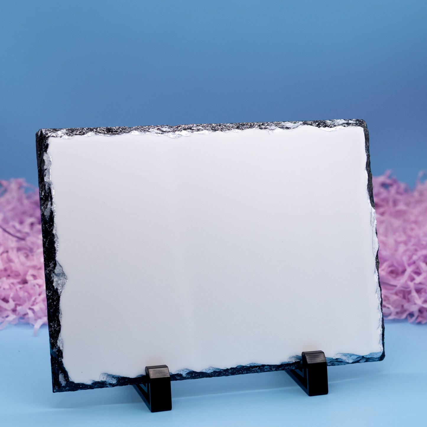 A rectangular plaque with a rough dark natural stone border, displayed on a small black stand against a blue background with pink decorative elements.