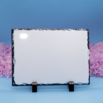 A rectangular slate with rough dark natural edges, displayed on a small black stand against a blue background with pink shredded paper accents.