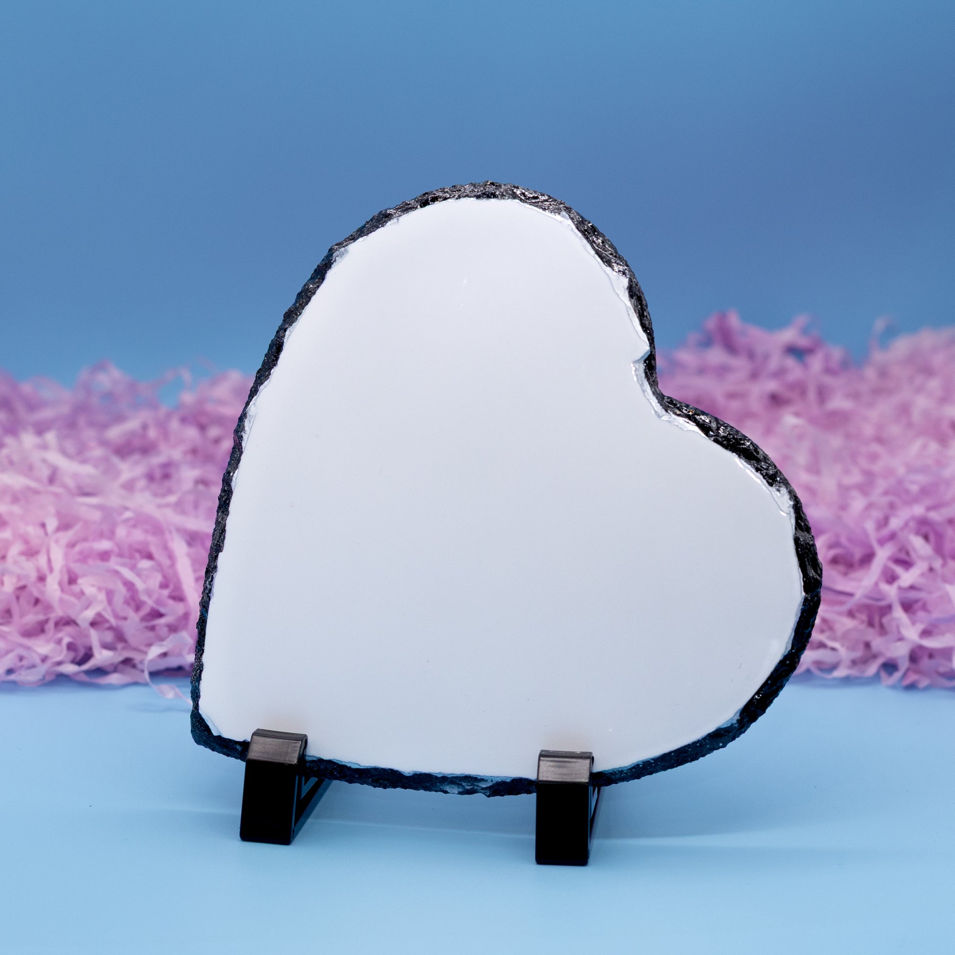 A heart-shaped polished plaque with a natural rough dark edge, displayed on a small black stand against a blue background with pink shredded paper.