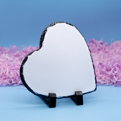 A heart-shaped polished stone plaque with a natural rough black edge, displayed on a small black stand against a blue background with pink shredded paper.