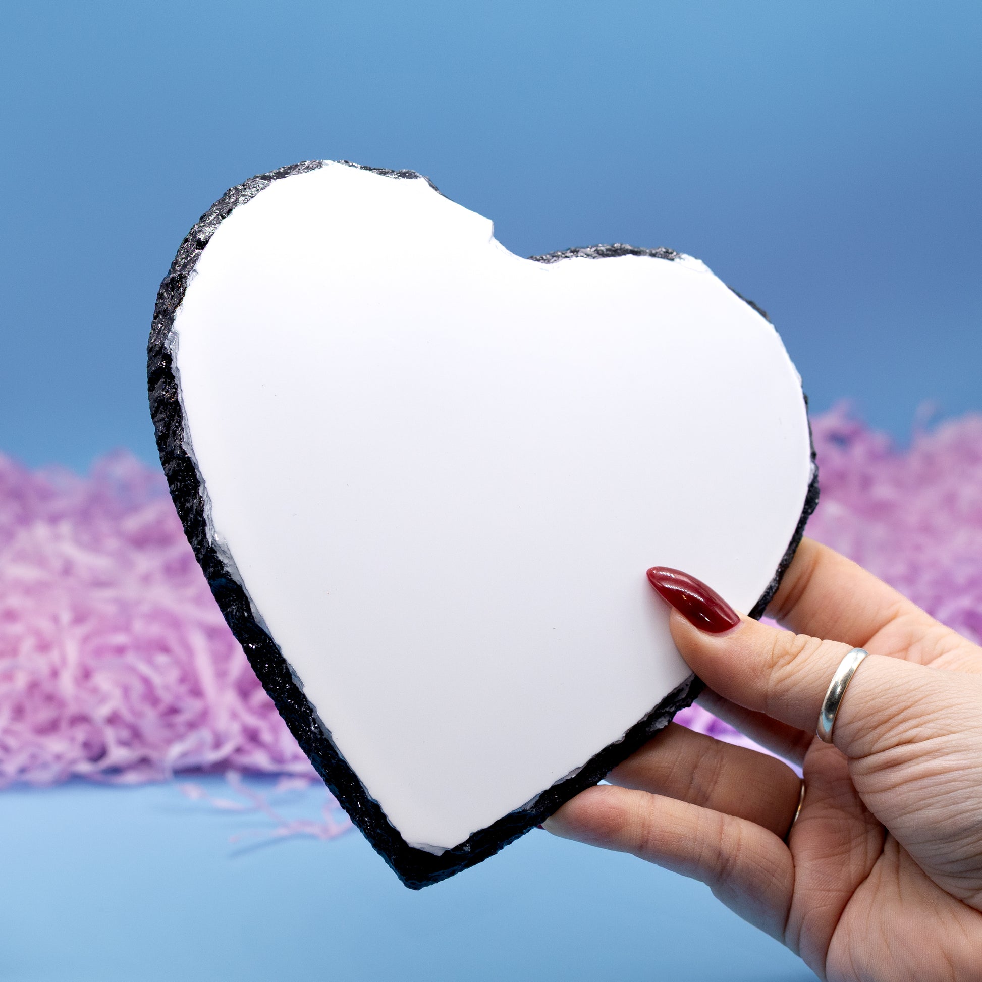A hand holds a heart-shaped slate with a black textured border, set against a blue background with pink shredded paper.