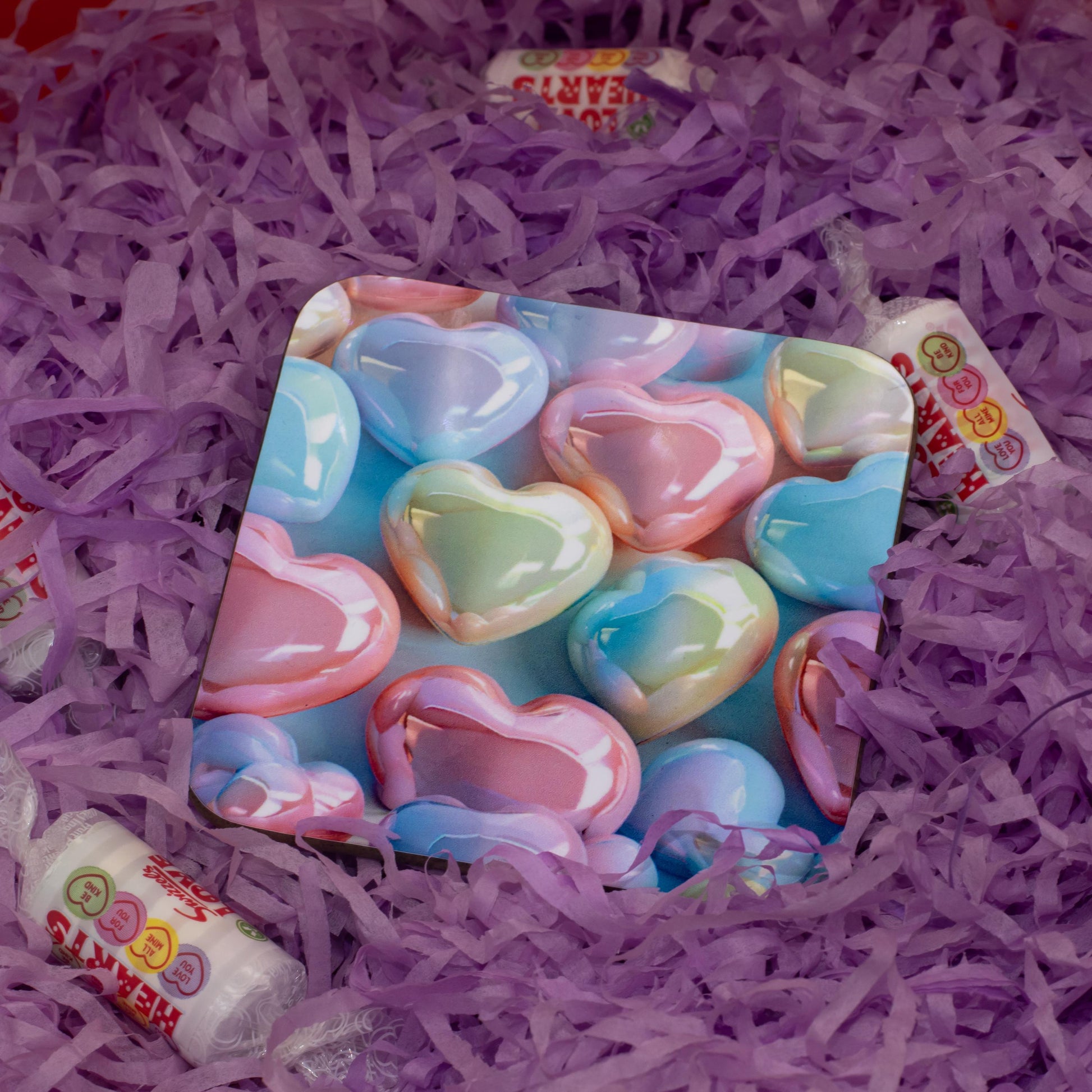 A square decorative coaster featuring a colorful candy heart pattern, surrounded by purple shredded paper and small wrapped candy rolls.