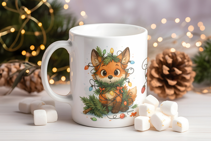 A white ceramic mug featuring a cute illustration of a smiling orange fox wearing a green pine wreath and colorful holiday lights, styled on a white surface with marshmallows and pinecones against a warm, blurred festive background.