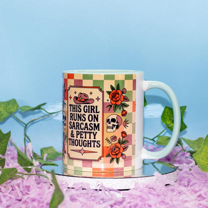 A white ceramic mug with a colorful retro checkered pattern and illustrations of a cowboy hat, roses, and a skull, featuring the text 'THIS GIRL RUNS ON SARCASM & PETTY THOUGHTS'. The mug is styled on a mirrored surface with purple shredded paper and green ivy against a light blue background.