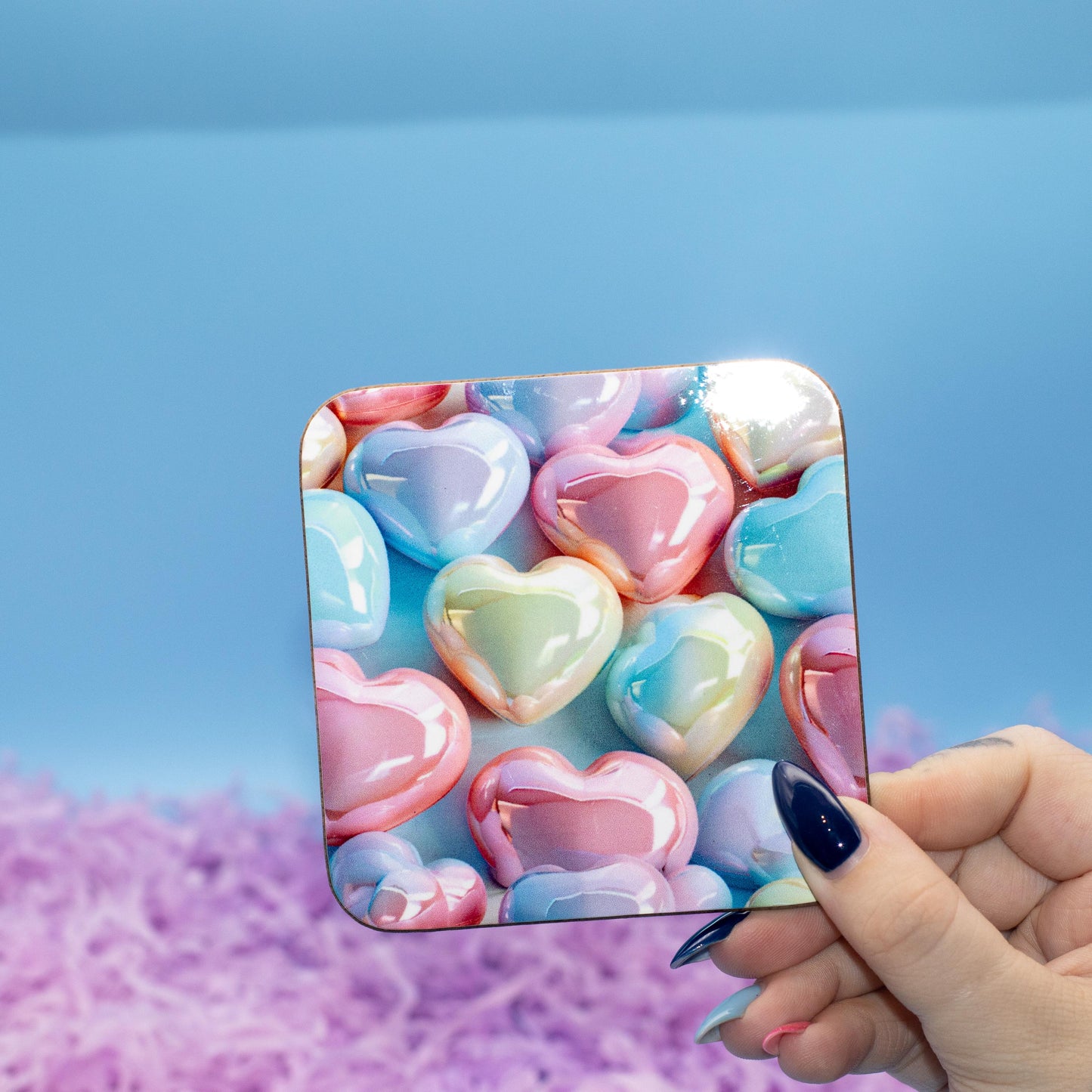Square hard backed coaster featuring a colourful pattern of glossy pastel heart candies, set against a blue background with purple shredded paper.