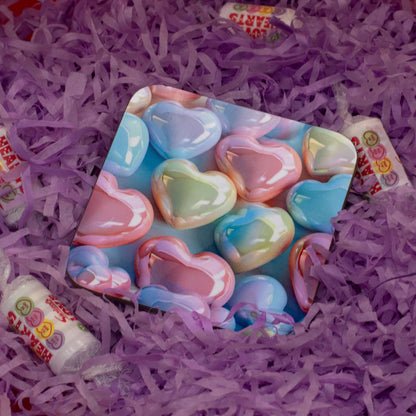 A square coaster featuring a pastel heart-shaped candy pattern, nestled in purple shredded paper with small wrapped candies around it.