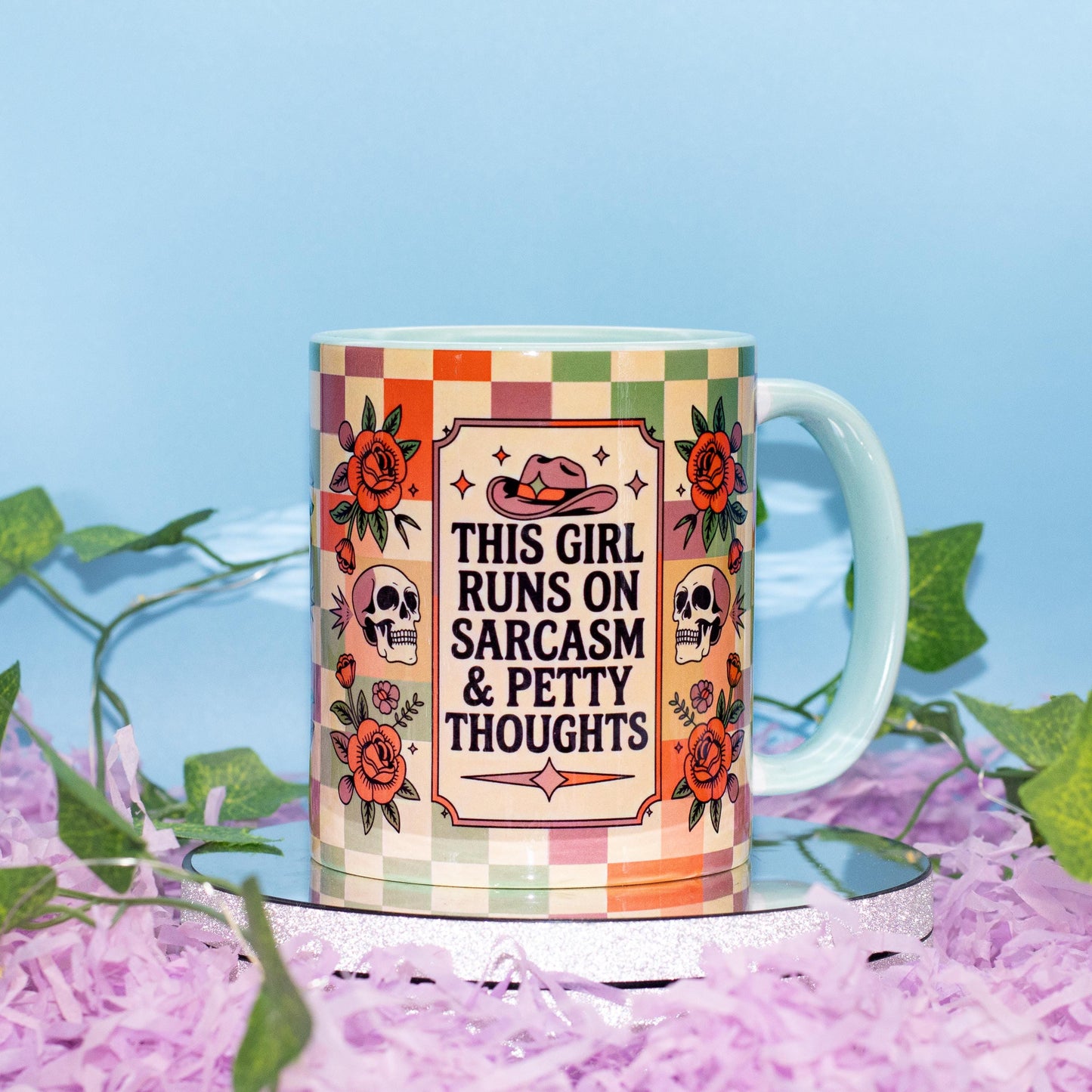 A ceramic mug with a colorful retro checkered pattern and floral illustrations, featuring the text 'THIS GIRL RUNS ON SARCASM & PETTY THOUGHTS' in the center. The mug is displayed on a reflective silver tray surrounded by purple shredded paper and green ivy against a light blue background.