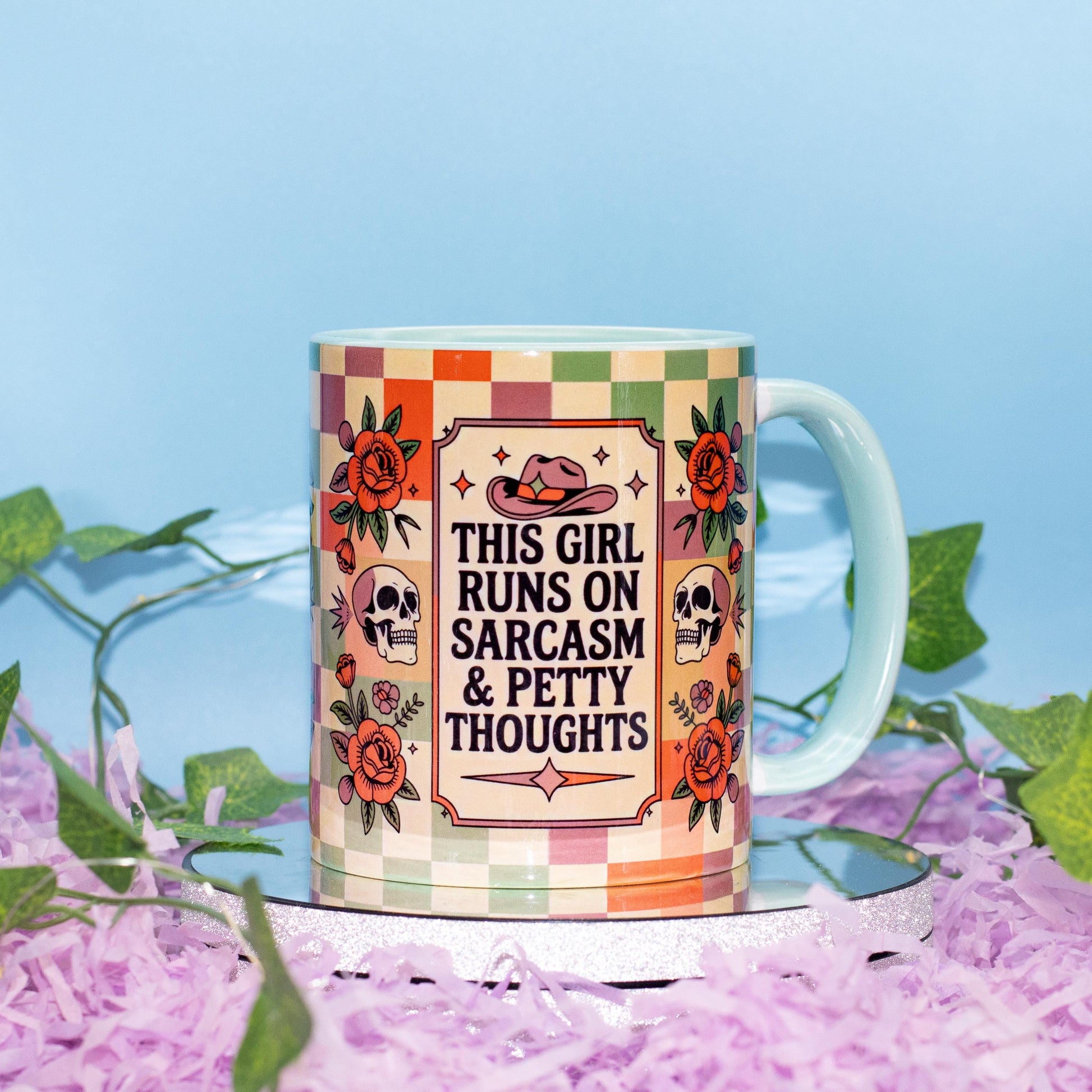 A ceramic mug with a colorful retro checkered pattern and floral illustrations, featuring the text 'THIS GIRL RUNS ON SARCASM & PETTY THOUGHTS' in the center. The mug is displayed on a reflective silver tray surrounded by purple shredded paper and green ivy against a light blue background.