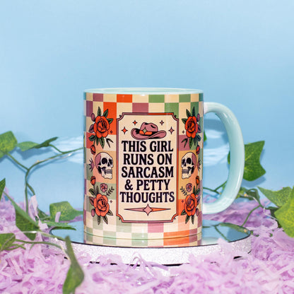 A ceramic mug with a colorful retro checkered pattern and floral illustrations, featuring the text 'THIS GIRL RUNS ON SARCASM & PETTY THOUGHTS' in the center. The mug is displayed on a reflective silver tray surrounded by purple shredded paper and green ivy against a light blue background.