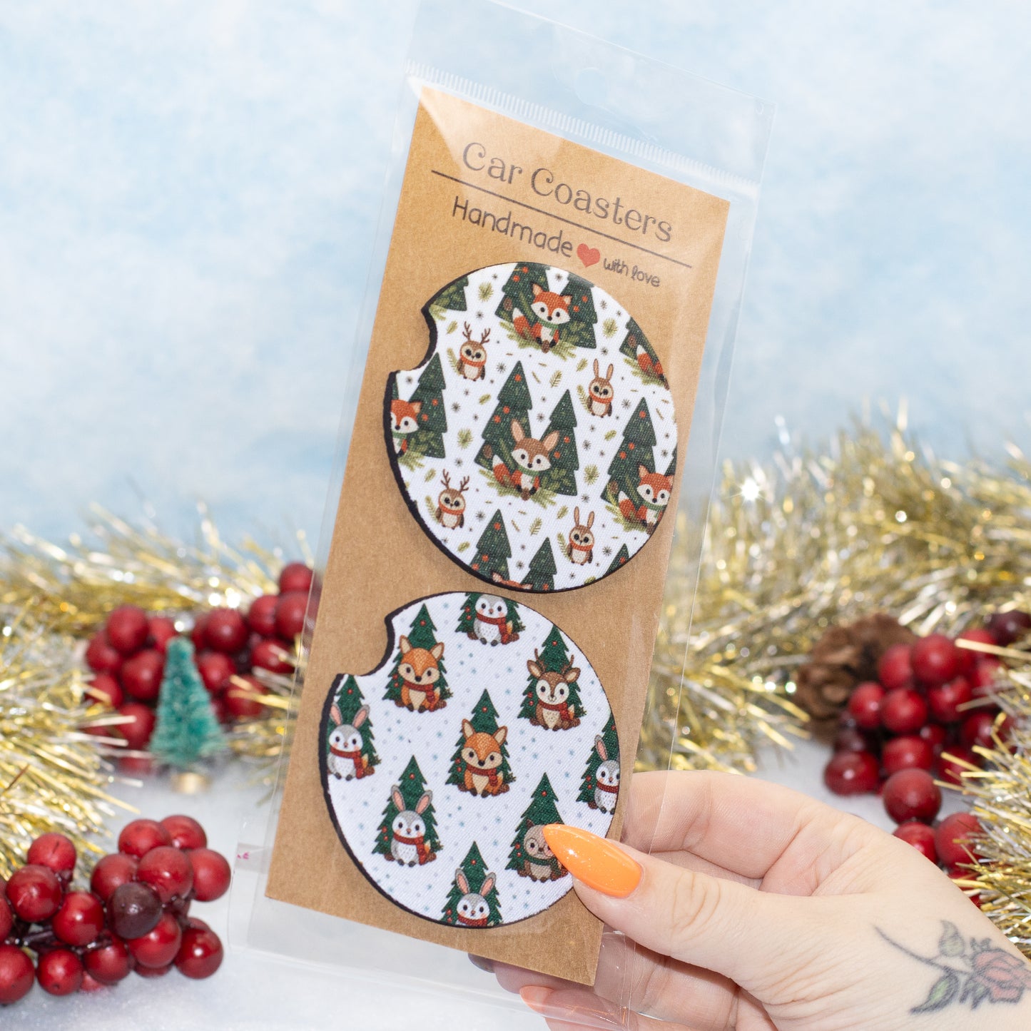 A hand holds a clear plastic package containing two car coasters with a festive woodland pattern of owls, Christmas trees, and pine cones on a white background. 