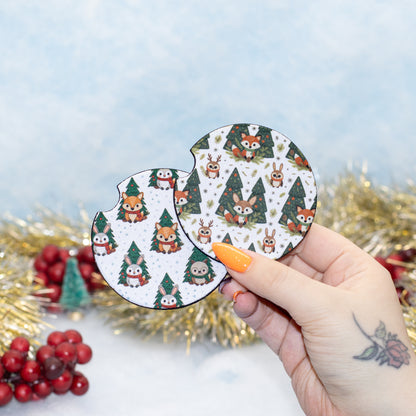 A hand holds two round coasters featuring a festive woodland pattern of foxes, rabbits, and Christmas trees. The background is decorated with gold tinsel and red berries against a soft, light blue textured backdrop.