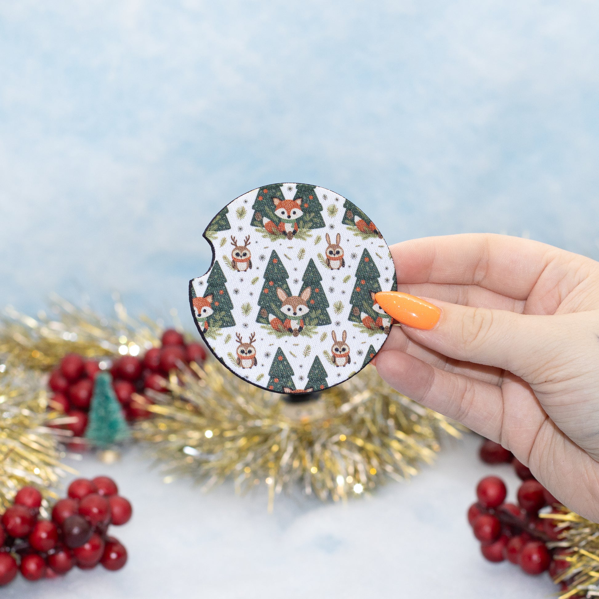 A hand holds a round coaster featuring a festive Christmas pattern of foxes, rabbits, and Christmas trees. The background is a soft light blue with blurred gold tinsel and red berries.