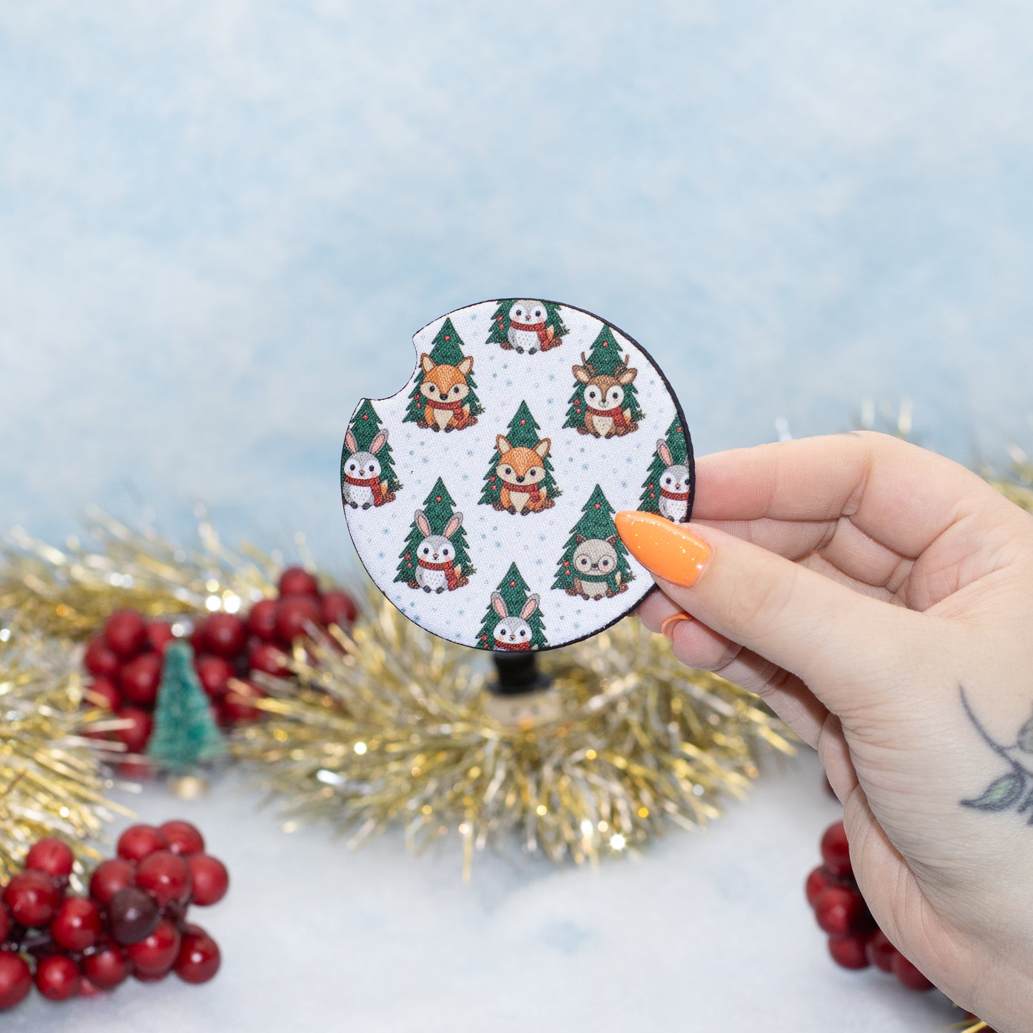 A hand holds a round coaster featuring a white background with a repeating Christmas tree and owl pattern. The background is a soft light blue textured surface decorated with gold tinsel and red berries.