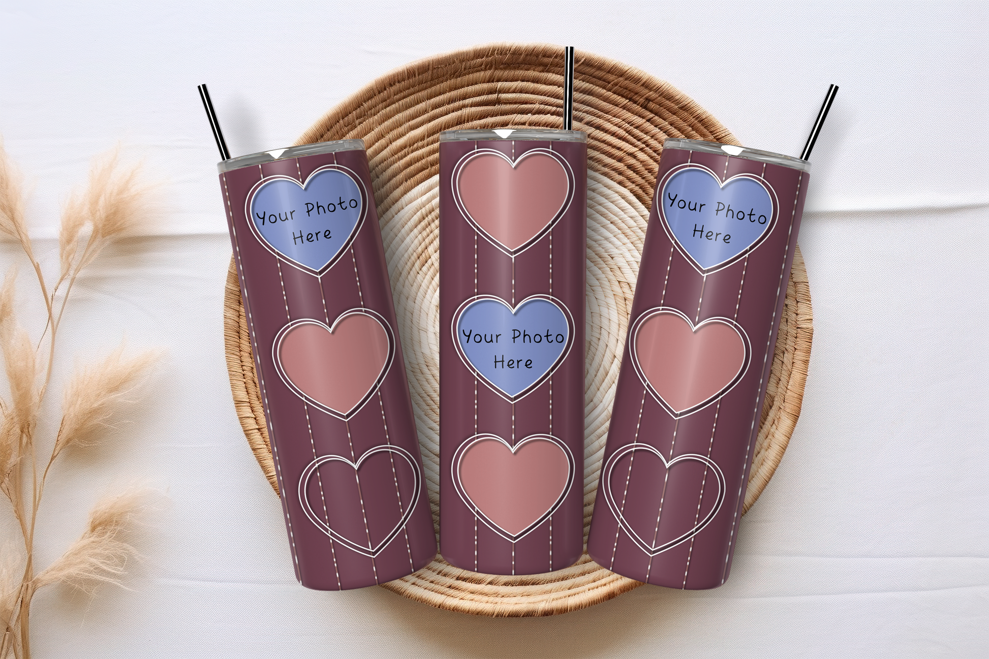Three insulated tumblers with metal straws, featuring a dark red background and heart graphics, arranged on a round woven tray against a white wood background.
