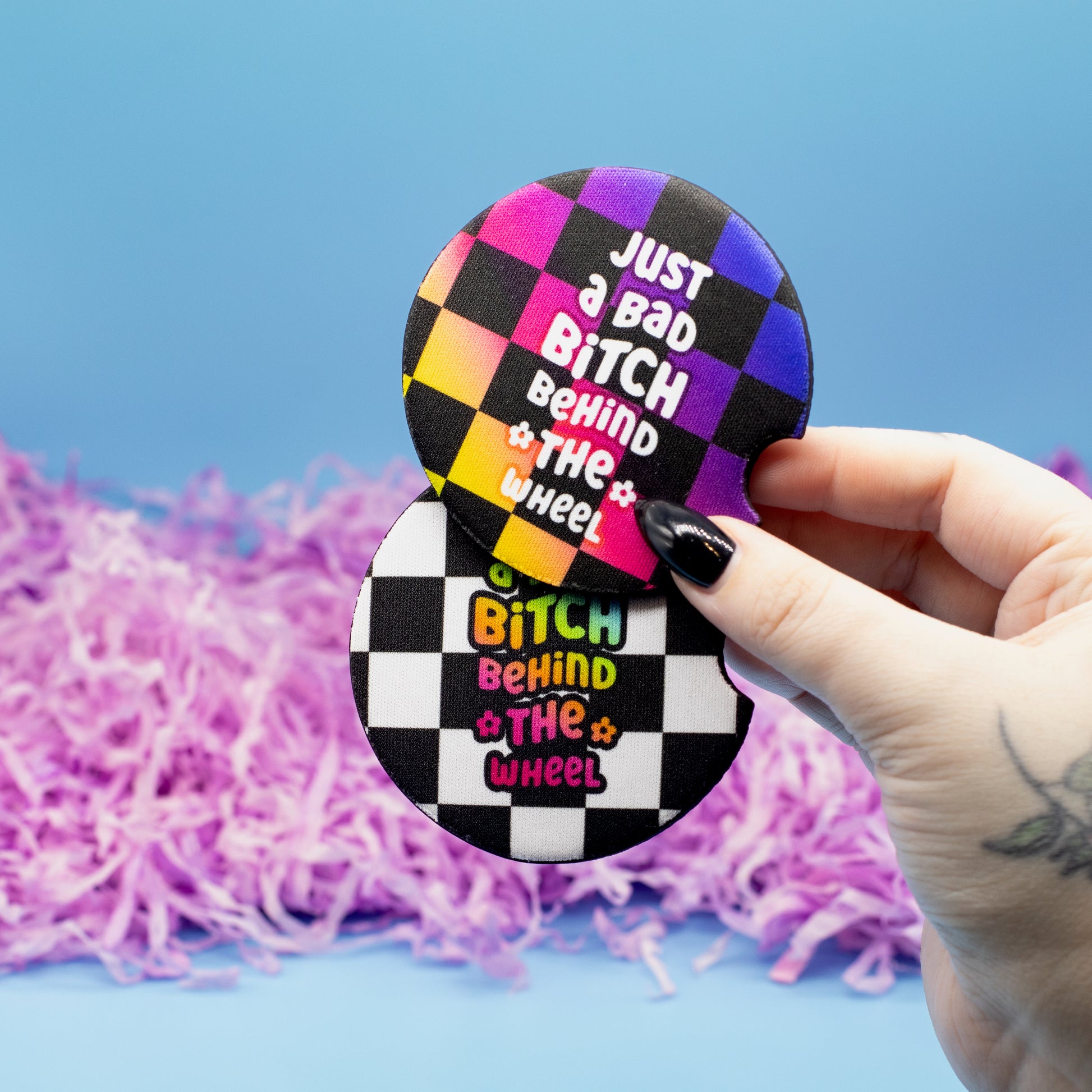 A hand holds two round car coasters featuring a black and white checkered wheel design with the text 'Just a Bad Bitch Behind the Wheel' in colourful lettering, set against a blue background with purple shredded paper.