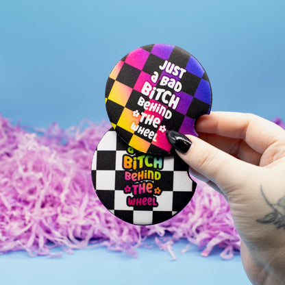 A hand holds two round car coasters featuring a black and white checkered wheel design with the text 'Just a Bad Bitch Behind the Wheel' in colourful lettering, set against a blue background with purple shredded paper.