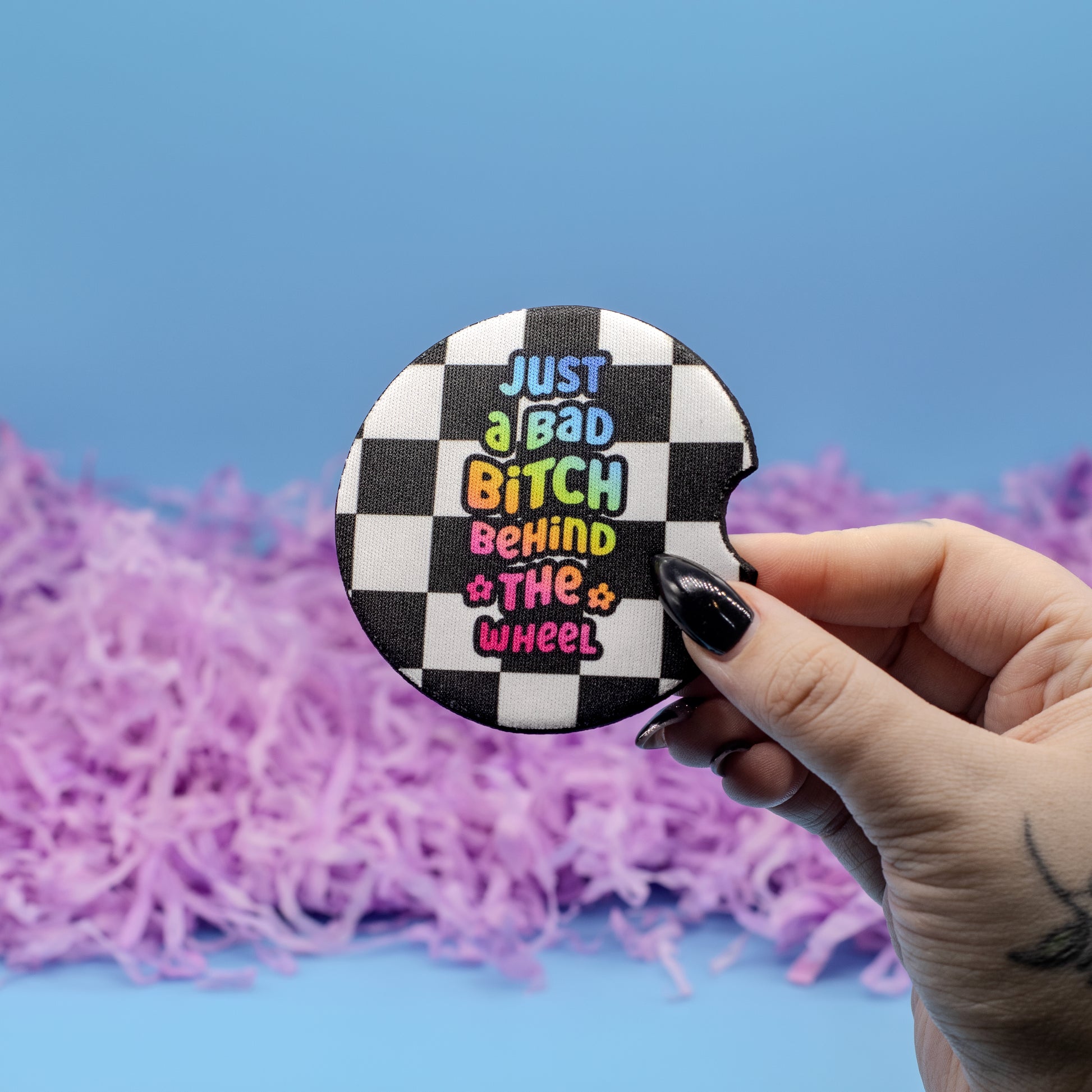 A hand holds a round black-and-white checkered coaster featuring the text 'JUST a BAD BITCH BEHIND THE WHEEL' in colourful bubble letters, against a blue background with purple shredded paper.