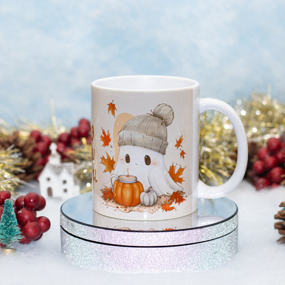 A white ceramic mug with a cute cartoon ghost wearing a gray beanie and holding an orange pumpkin, surrounded by autumn leaves, displayed on a round silver glitter pedestal with festive holiday decorations in the background.