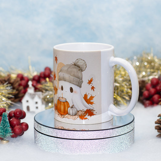 A white ceramic mug with a cute cartoon ghost wearing a gray beanie and holding an orange pumpkin, displayed on a round silver glitter stand with seasonal holiday decorations in the background.