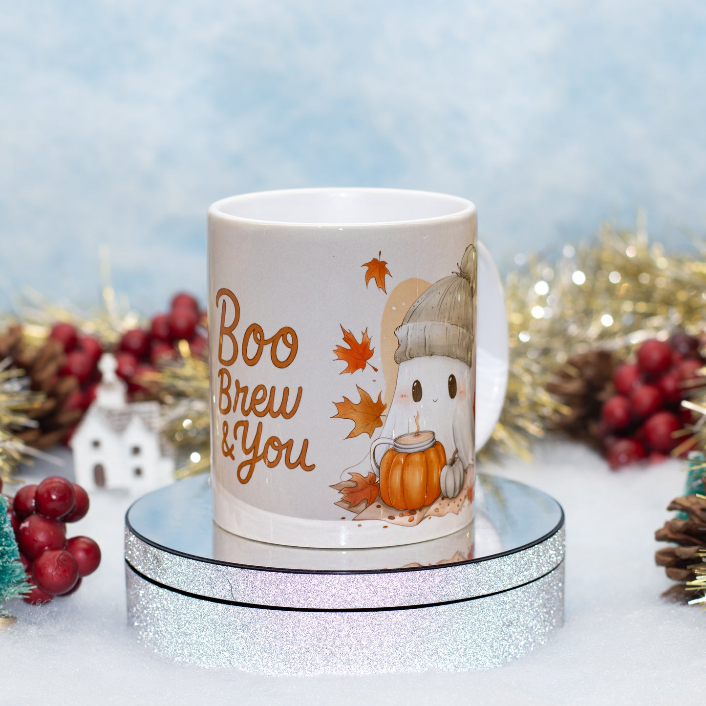 A white ceramic mug with a cute cartoon ghost wearing a hat and holding a pumpkin, surrounded by autumn leaves and the text 'Boo Brew & You'. The mug is displayed on a round silver glitter stand against a festive background with gold tinsel, pinecones, and red berries.