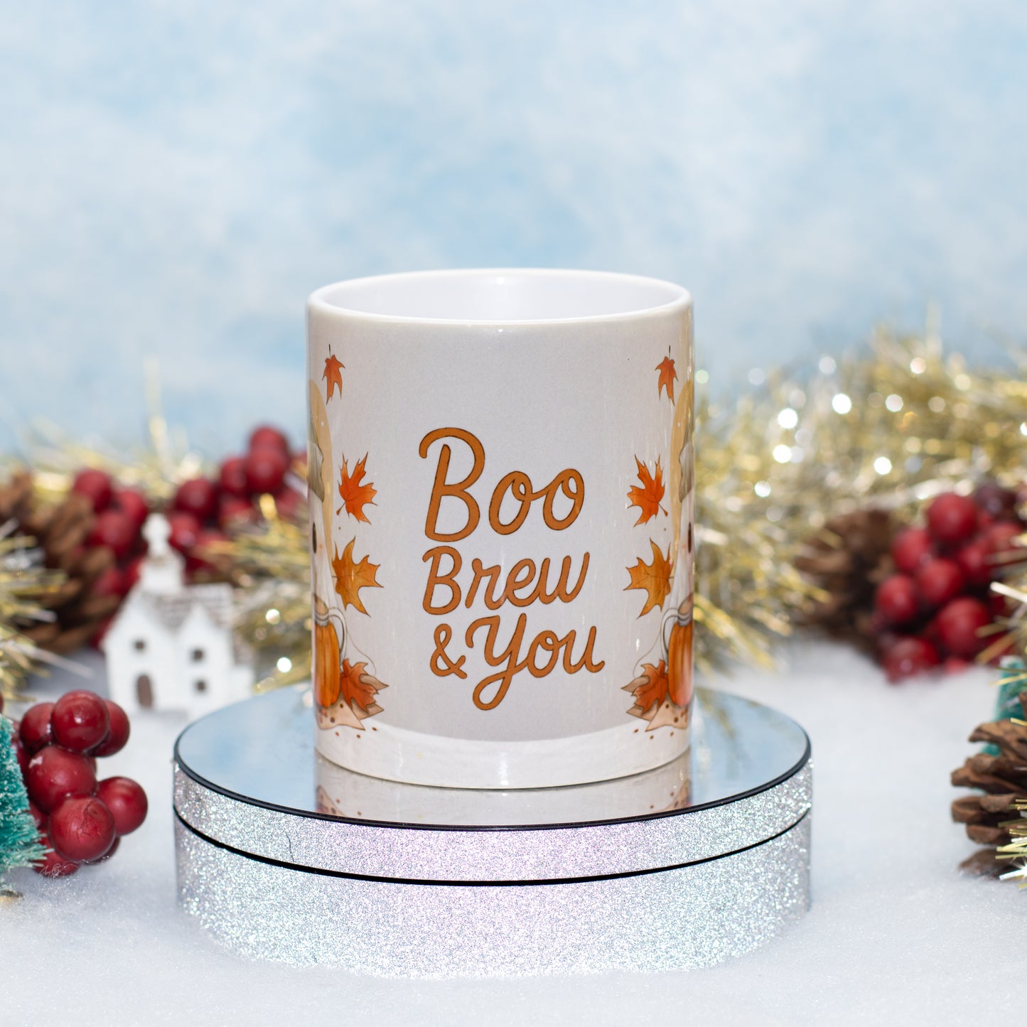A white ceramic mug with 'Boo Brew & You' printed in orange script, surrounded by orange autumn leaves, displayed on a round silver glitter pedestal with festive holiday decorations in the background.