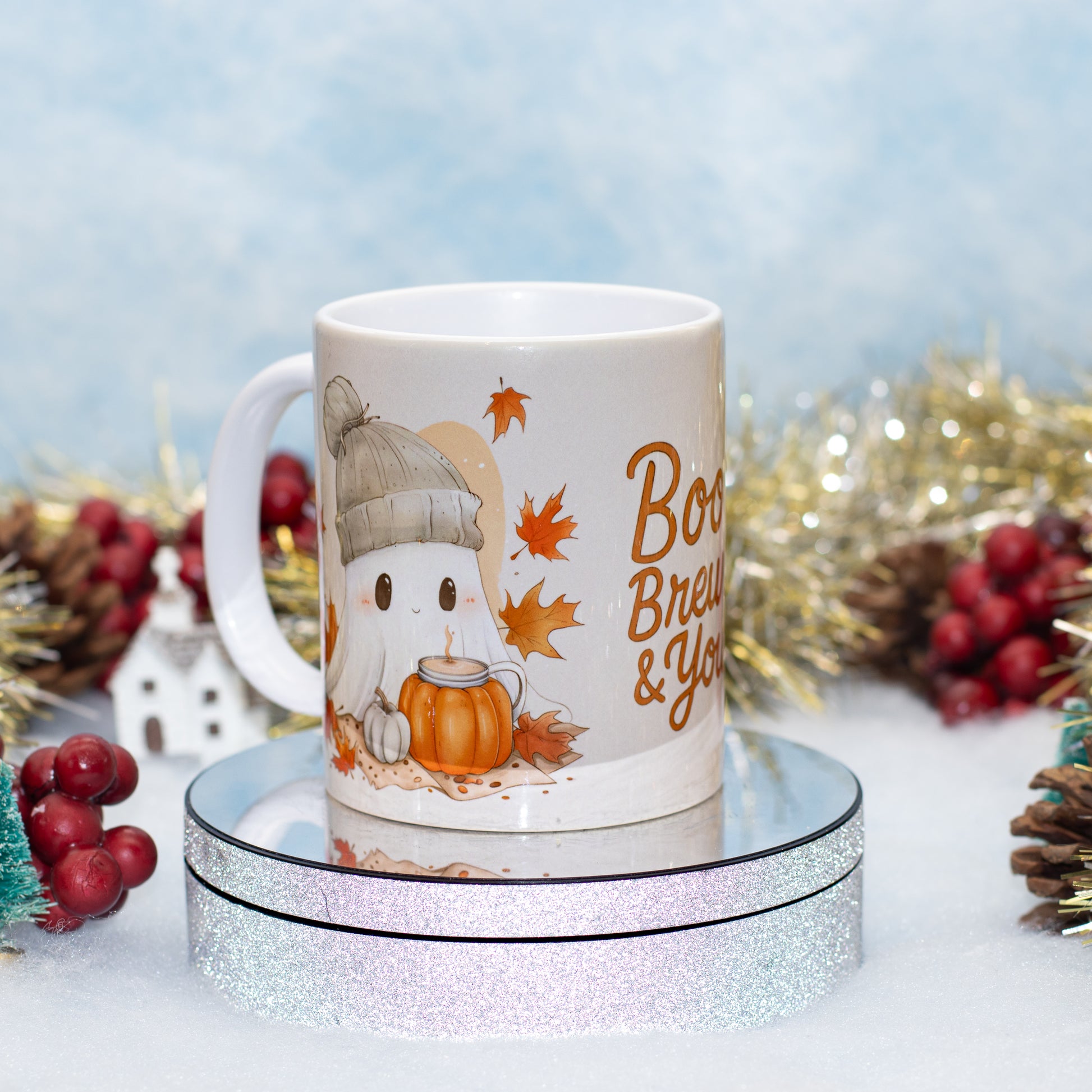 A white ceramic mug with a cute cartoon ghost wearing a beanie and holding a pumpkin, surrounded by autumn leaves and the text 'Boo Brew & Y'all'. The mug is displayed on a round glittery stand with festive holiday decorations in the background.