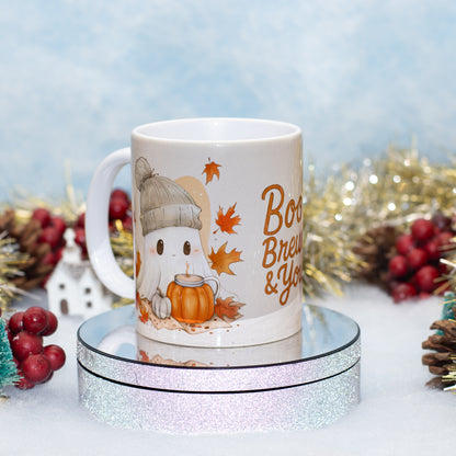 A white ceramic mug with a cute cartoon ghost wearing a beanie and holding a pumpkin, surrounded by autumn leaves and the text 'Boo Brew & Y'all'. The mug is displayed on a round glittery stand with festive holiday decorations in the background.