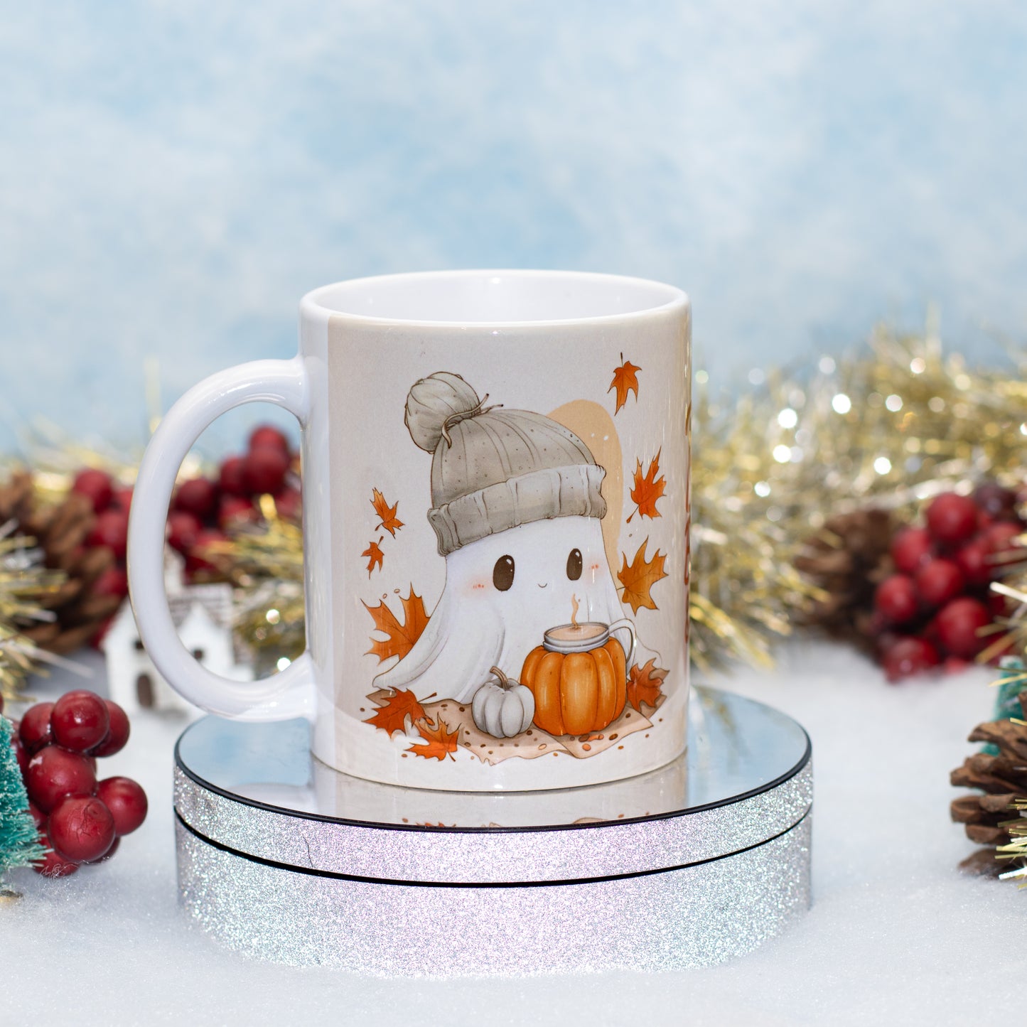 A white ceramic mug with a cute cartoon ghost wearing a gray beanie and sitting beside a small orange pumpkin, surrounded by autumn leaves. The mug is displayed on a round silver glitter pedestal with festive holiday decorations in the background.