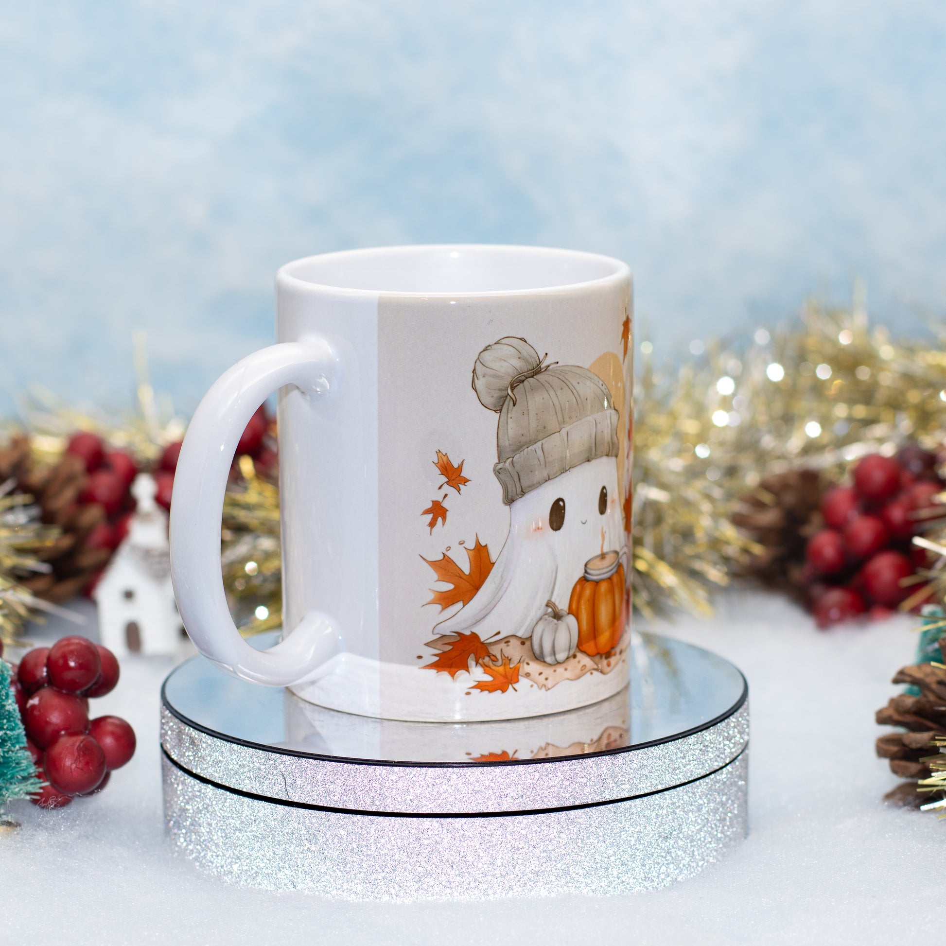 A white ceramic mug with a cute ghost illustration wearing a gray beanie and holding an orange pumpkin, styled on a mirrored stand with autumn decorations in the background.