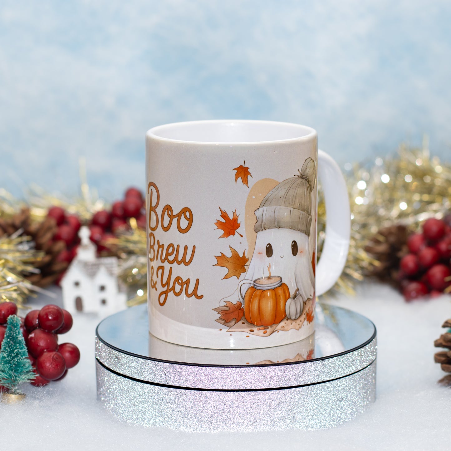 A white ceramic mug with a cute cartoon ghost wearing a beanie and holding a pumpkin, surrounded by autumn leaves and the words 'Boo Brew You'. The mug is displayed on a round silver glitter pedestal with festive holiday decorations in the background.